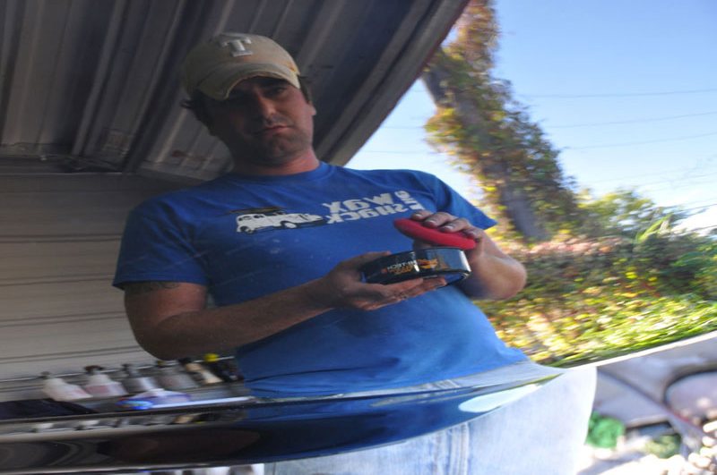 A Wax Shack technician hand-waxing the roof of a dark blue car, with trees reflected in the paint.
