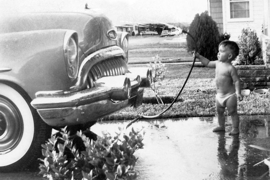 Black and white photograph of a young boy in shorts hosing down a 1950s car in a driveway.