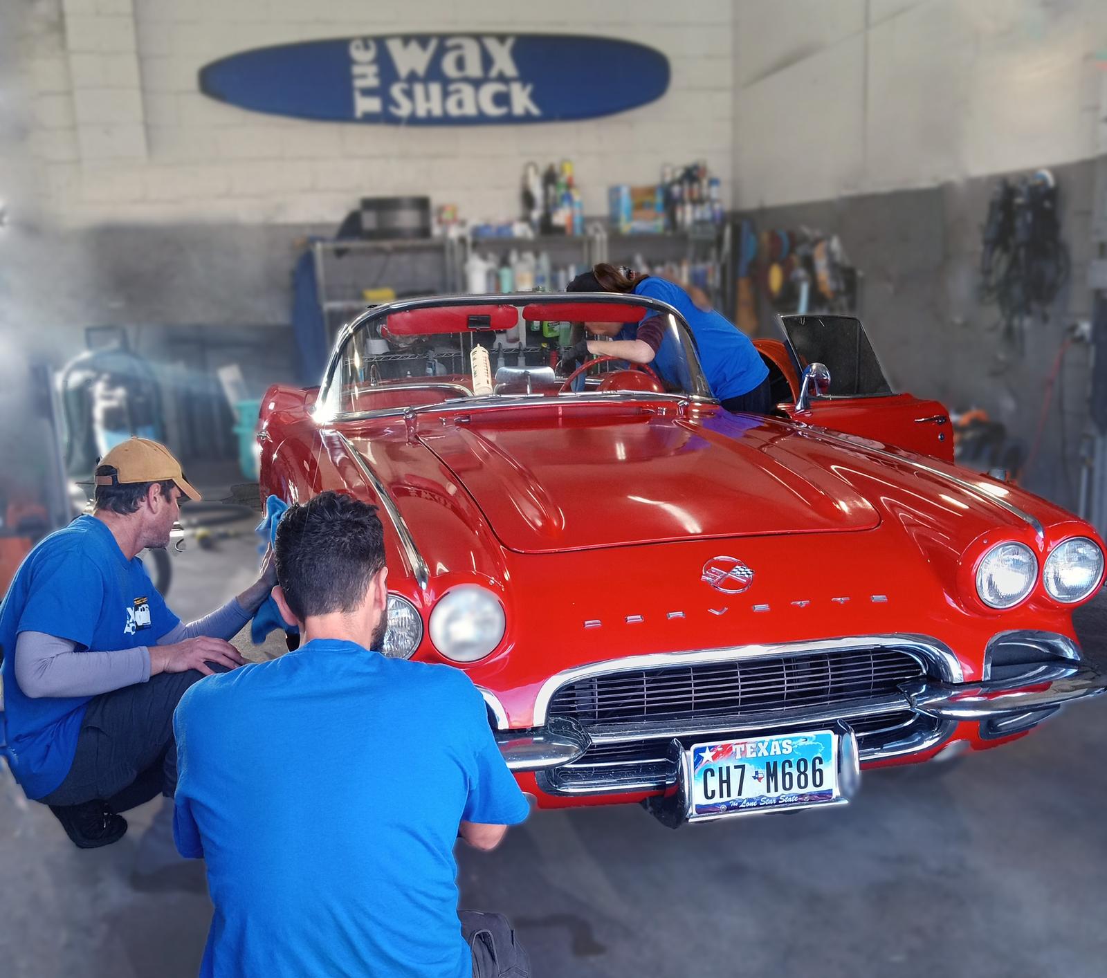 Three Wax Shack detailers in blue shirts working on a red 1962 Corvette convertible inside the Austin shop, beneath the Wax Shack sign.