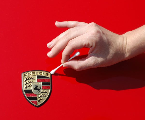 A hand using a cotton swab to clean around the raised crest badge on a red car’s bonnet.